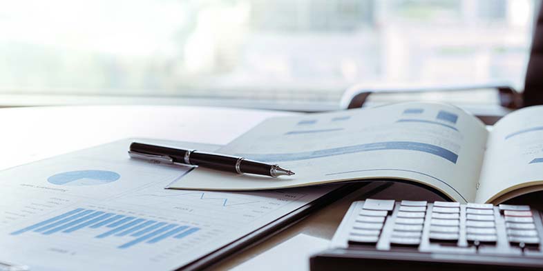 Pen resting on an open book full of charts and graphs. A calculator is also visible just in the corner of the frame.